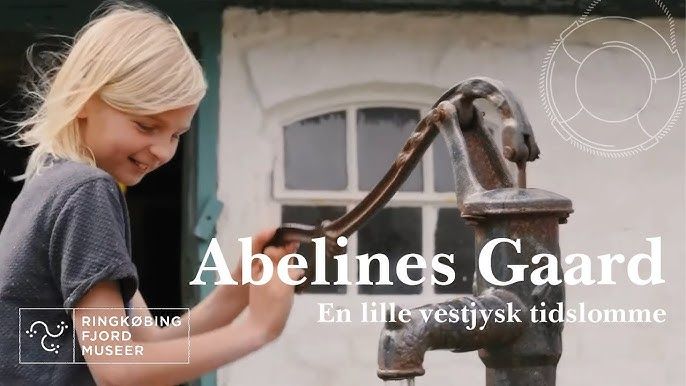 Smilende barn pumper vand ved en gammel pumpe, foran hvid bygning med vindue.